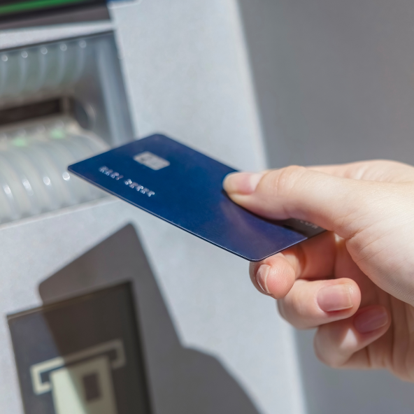 Kobieca dłoń trzyma w ręku niebieską kartę płatniczą i zbliża ją do bankomatu