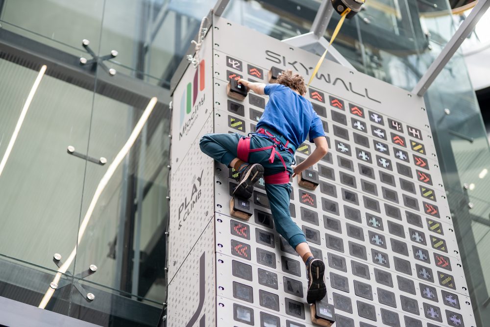Zdjęcie przedstawia osobę w uprzęży asekuracyjnej, która wspina się po nowoczesnej, pionowej ściance wspinaczkowej o nazwie „Skywall” z interaktywnymi, podświetlanymi panelami LED. Konstrukcja z logiem Galerii Młociny znajduje się wewnątrz nowoczesnego budynku ze szklanymi elementami architektonicznymi.