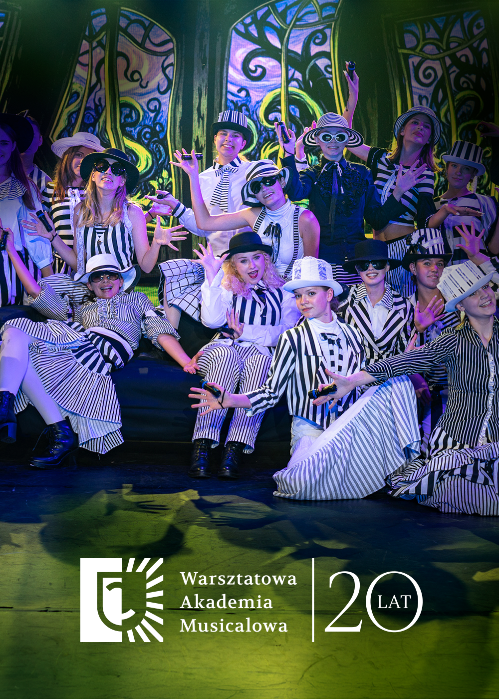 Grupa młodych artystów w czarno-białych, pasiakowych strojach i kapeluszach pozuje na scenie podczas występu teatralnego. W dolnej części widoczne jest logo „Warsztatowa Akademia Musicalowa” oraz rocznicowy napis „20 lat”.