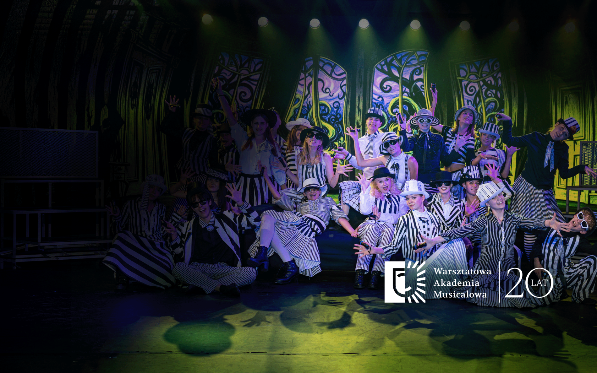 Grupa młodych artystów w czarno-białych, pasiakowych strojach i kapeluszach pozuje na scenie podczas występu teatralnego. W dolnej części widoczne jest logo „Warsztatowa Akademia Musicalowa” oraz rocznicowy napis „20 lat”.