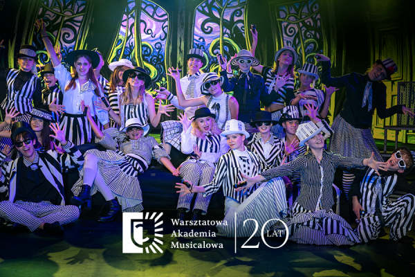 Grupa młodych artystów w czarno-białych, pasiakowych strojach i kapeluszach pozuje na scenie podczas występu teatralnego. W dolnej części widoczne jest logo „Warsztatowa Akademia Musicalowa” oraz rocznicowy napis „20 lat”.