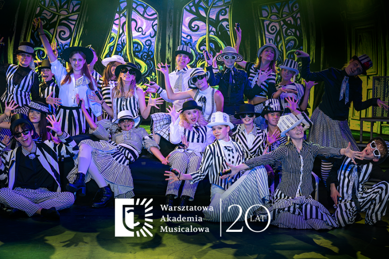 Grupa młodych artystów w czarno-białych, pasiakowych strojach i kapeluszach pozuje na scenie podczas występu teatralnego. W dolnej części widoczne jest logo „Warsztatowa Akademia Musicalowa” oraz rocznicowy napis „20 lat”.