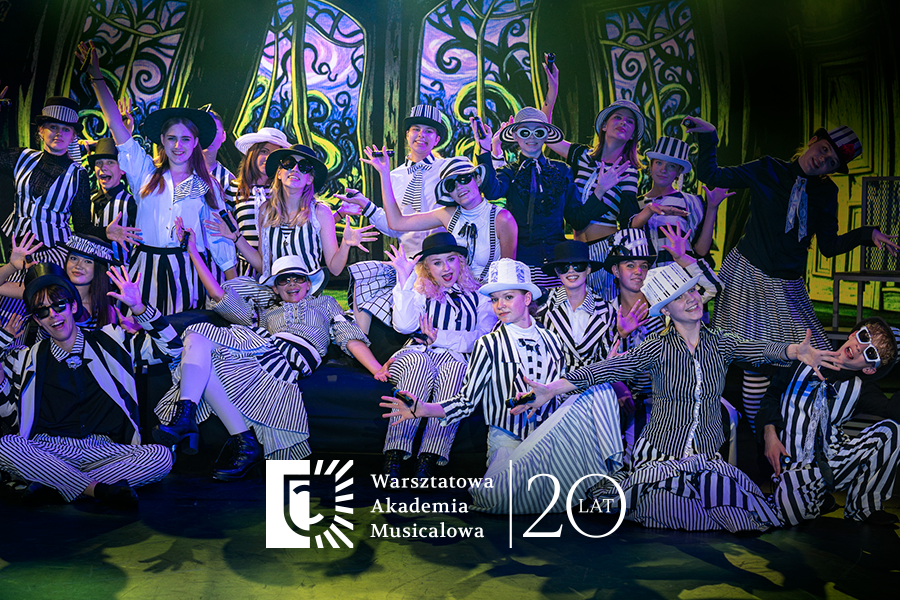 Grupa młodych artystów w czarno-białych, pasiakowych strojach i kapeluszach pozuje na scenie podczas występu teatralnego. W dolnej części widoczne jest logo „Warsztatowa Akademia Musicalowa” oraz rocznicowy napis „20 lat”.