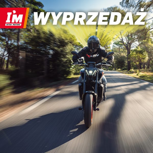 Motocyklista jadący po asfaltowej drodze w kasku i kurtce motocyklowej na tle zielonych drzew z czerwoną identyfikacją Inter Motors i napisem Wyprzedaż w Galerii Młociny w Warszawie
