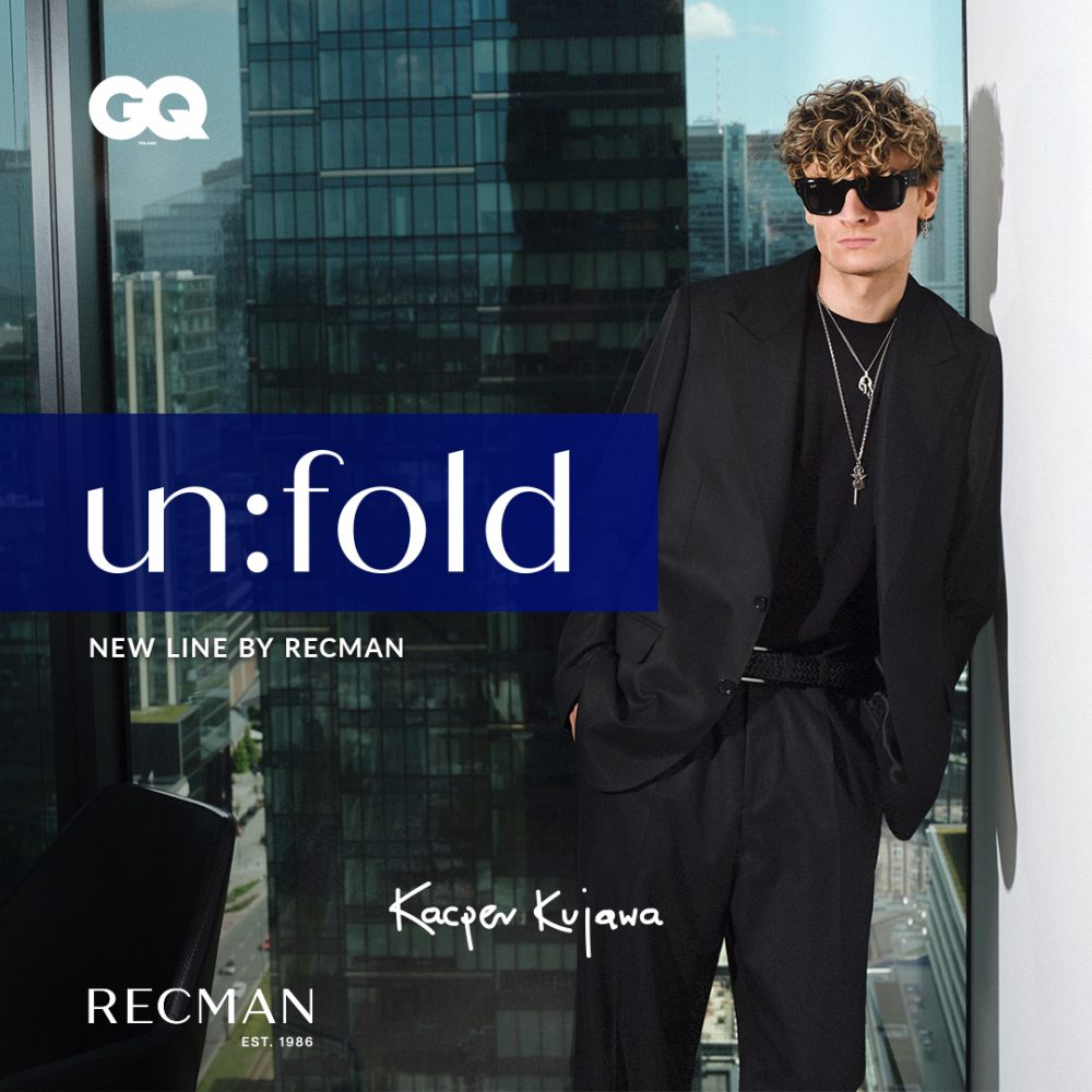 Elegancka grafika promocyjna nowej linii „un:fold” marki Recman z udziałem Kacpra Kujawy. Na pierwszym planie widoczny jest mężczyzna w czarnym garniturze, ciemnej koszulce, okulary przeciwsłonecznych i biżuterii, pozujący na tle okna z panoramą miejskich wieżowców. Po lewej stronie znajduje się niebieski element z napisem „un:fold NEW LINE BY RECMAN”, a w rogach grafiki umieszczono logotypy magazynu GQ oraz marki Recman.