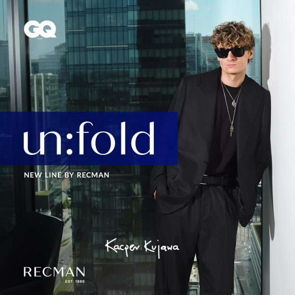 Elegancka grafika promocyjna nowej linii „un:fold” marki Recman z udziałem Kacpra Kujawy. Na pierwszym planie widoczny jest mężczyzna w czarnym garniturze, ciemnej koszulce, okulary przeciwsłonecznych i biżuterii, pozujący na tle okna z panoramą miejskich wieżowców. Po lewej stronie znajduje się niebieski element z napisem „un:fold NEW LINE BY RECMAN”, a w rogach grafiki umieszczono logotypy magazynu GQ oraz marki Recman.